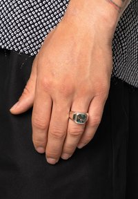 Anillo hexagonal de plata con un incrustación iridiscente, llevado en el dedo índice, con una mano texturizada contra un fondo de tela oscura.