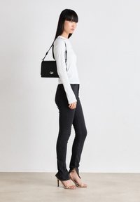 Svart rektangulær håndveske med logo, båret over skulderen. Modell i tettsittende svarte jeans og hvit langermet topp, høyhælte sandaler.