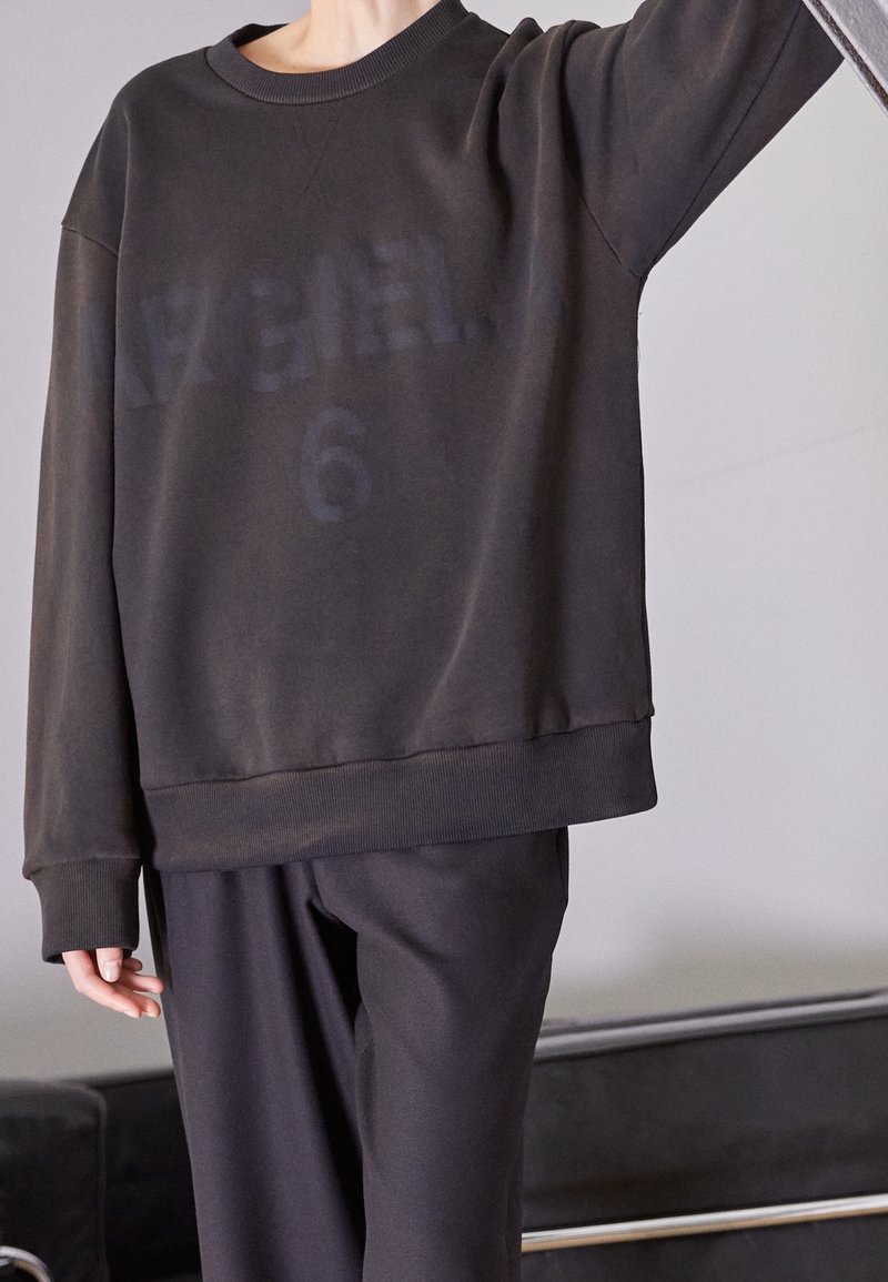 Schwarzer Sweatshirt mit rundem Halsausschnitt, das einen verblassten Aufdruck "ARGIELA 6" auf der Vorderseite und gerippte Bündchen aufweist. Kombiniert mit dunklen Hosen.