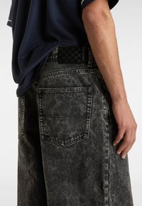 Sorte denimshorts med en falmet tekstur og en ternet patch på taljen. Baglommen er synlig på venstre side.
