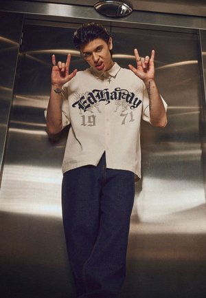 Jeune homme portant une chemise beige Ed Hardy et un pantalon sombre faisant des signes de la main rock devant une paroi métallique d'ascenseur.