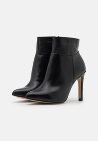 Botas de tobillo de cuero negro con punta afilada y tacón stiletto alto. Presentan una textura suave y un diseño de caña cónica.