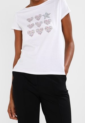 Wit T-shirt met korte mouwen met negen bloemvormen—acht harten en één ster—gedragen met een zwarte broek door een staand persoon.