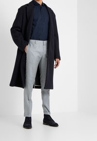 Un abrigo de lana azul marino, una camisa de botones azul oscuro, pantalones ajustados de cuadros claros y zapatos de ante azul marino crean un atuendo moderno y coordinado.