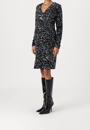 Femme portant une robe noire pailletée longueur genou avec des manches longues et un col en V, associée à des bottes noires à talons hauts, pointues et montant jusqu'au genou.