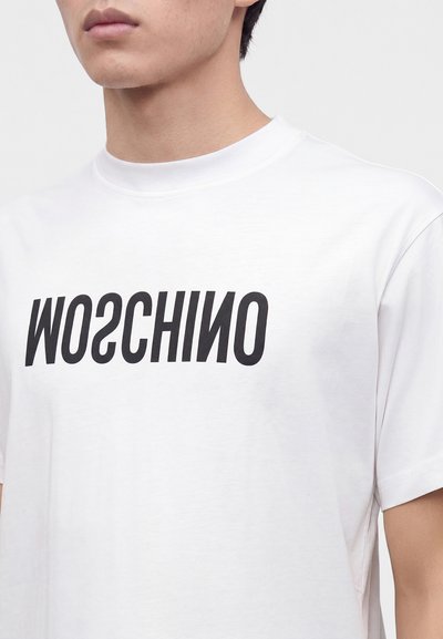 Ένα άτομο φοράει ένα λευκό T-shirt με τη λέξη MOSCHINO τυπωμένη με μεγάλα μαύρα γράμματα στο στήθος.