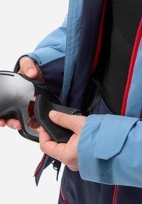 Ski goggles held in a hand, featuring a mirrored lens and black foam padding. The hand is wearing a blue and navy jacket with red accents.