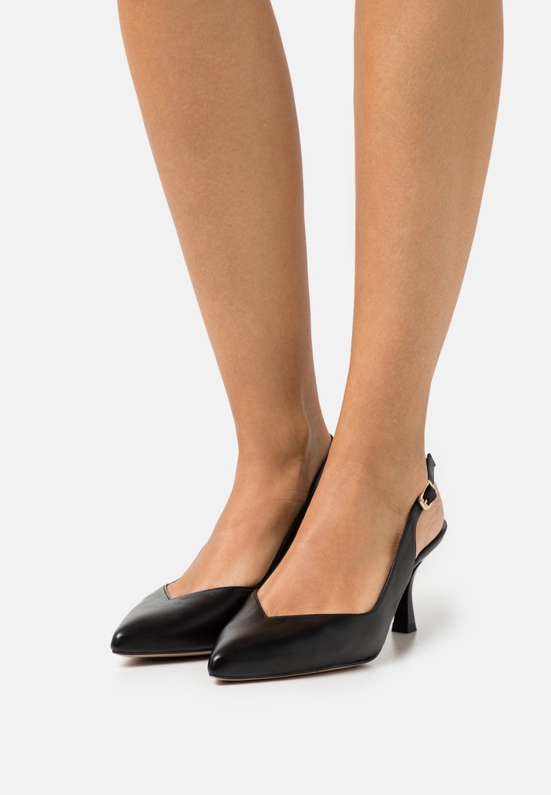 Jambes portant des talons slingback noirs à bout pointu avec un talon stiletto moyen, montrant la peau au-dessus de la cheville sur un fond uni.