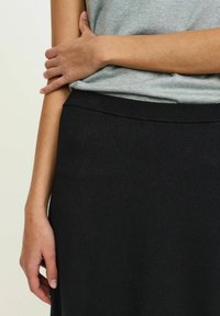 Čierna pletená sukňa s hladkou textúrou, vybavená širokým elastickým pásom. Ruka modelky spočíva na páse a predvádza strih odevu.