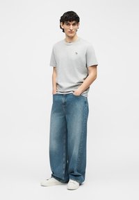 Νέος άνδρας στέκεται με τα χέρια στις τσέπες, φοράει ανοιχτό γκρι κοντομάνικο πουκάμισο, φαρδιά μπλε τζιν παντελόνια και λευκά αθλητικά παπούτσια σε λευκό φόντο.