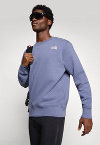 Mann med store svarte solbriller, blå The North Face hettegenser, svarte bukser, holder en svart ryggsekkstropp over den ene skulderen.
