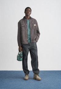 Šedá džínová bunda s předními kapsami, zelená košile, tmavé džíny, světle béžové boty a malá tyrkysová taška. Jednoduchý design a neutrální barvy.