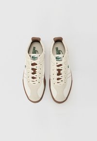 Zapatillas Lacoste en lona color blanco roto con detalles en marrón. Cuentan con cordones redondos, un logo verde y suelas de goma. Diseño atlético clásico.