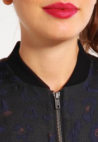 Svart bomberjacka med texturerat tyg, med dragkedjestängning och ribbad krage. Materialet har ett subtilt mönster i mörkblått och vinröd.