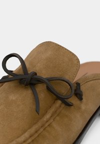 Καφέ μοκασίνι από σουέτ με στρογγυλερή μύτη, διακοσμητικό φιόγκο από μαύρο δερμάτινο κορδόνι και αντίθετη ραφή κατά μήκος της εξωτερικής άκρης.