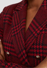 Rot-schwarz kariertes Blazer mit strukturiertem Stoff, doppelreihigem Design und Messingknöpfen auf der Vorderseite.