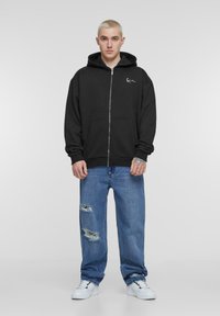 Felpa nera con zip, tasca frontale e logo sul petto, abbinata a jeans chiari strappati e sneakers bianche.