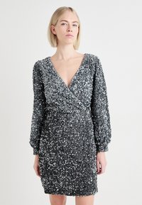 Vestido curto prateado coberto de lantejoulas, com mangas longas, decote em V e design envolvente. A superfície texturizada reflete a luz, criando um brilho.