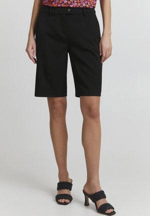Shorts bermuda noirs avec une coupe ajustée, dotés d'une fermeture à bouton et de passants de ceinture, assortis à des sandales à talons tissées noires.