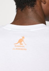 Biała koszulka z okrągłym dekoltem, z pomarańczowym logo kangura oraz napisem „KANGOL J.LINDEBERG” nadrukowanym z tyłu.