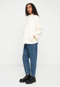 Donna con capelli ricci che indossa un pullover di pile crema, jeans blu e stivali con plateau neri, in piedi contro uno sfondo bianco uniforme.
