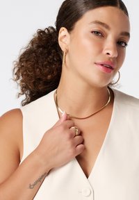 Gouden oorbellen in de vorm van een ring, een gladde gouden ketting en een ring; het model draagt een lichtbeige mouwloos topje met een v-hals en knopen.