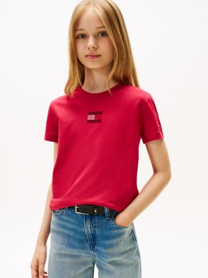 Rode katoenen T-shirt met een klein paillettenlogo op de voorkant, gecombineerd met lichtblauwe jeans en een zwarte riem.