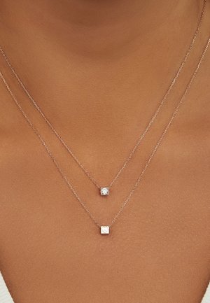 Colliers superposés avec des chaînes en argent ornées de deux pendentifs carrés ; l'un avec une pierre transparente, l'autre gravé d'un symbole de cœur.