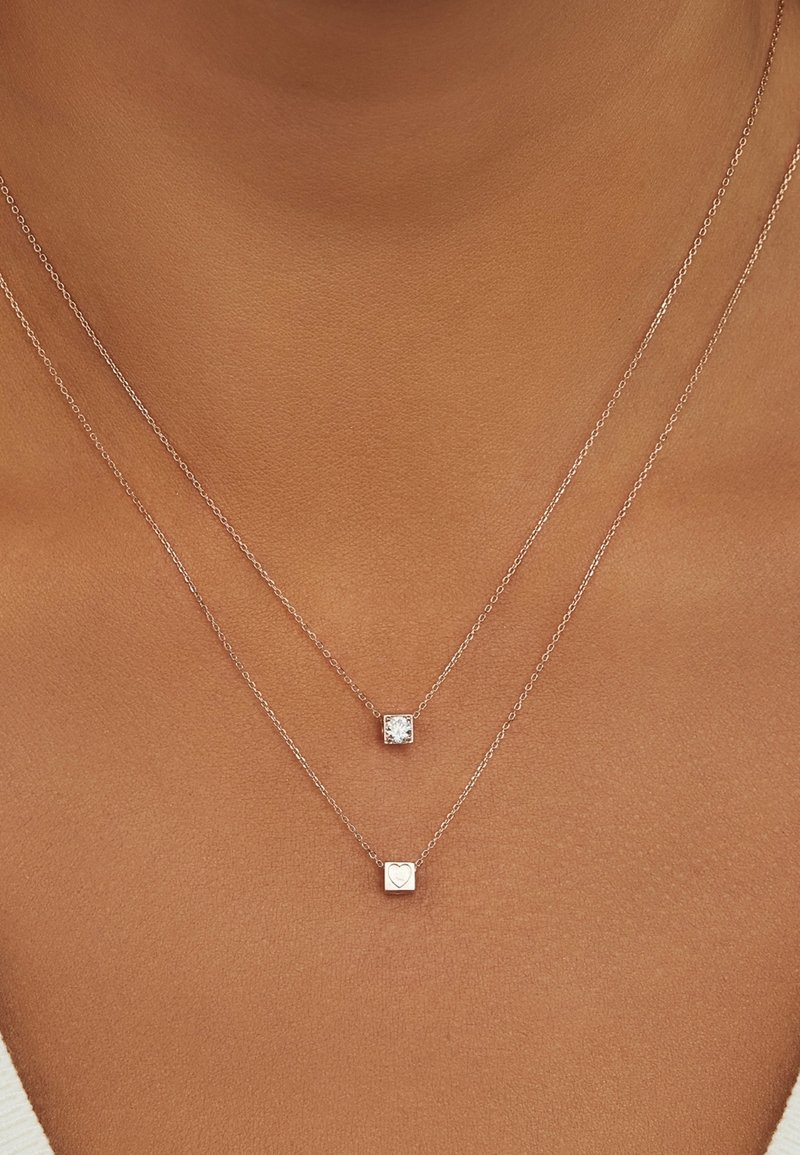 Colliers superposés avec des chaînes en argent ornées de deux pendentifs carrés ; l'un avec une pierre transparente, l'autre gravé d'un symbole de cœur.