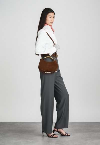 Brown suede handbag with a curved shape, gold hardware, shoulder strap, and a soft texture. Model wears gray trousers and white blouse.
