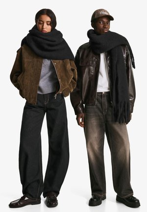 Femme portant une veste en daim marron, un pull gris et une écharpe noire. Homme en veste en cuir marron, t-shirt blanc, écharpe noire et jeans foncés.