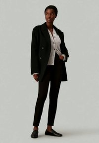 Mustmust kahe kätisega jope heledat halli kardigani peal, kombineeritud mustade kitsaste pükste ja mustade libisevate kingadega. Ükslane taust.