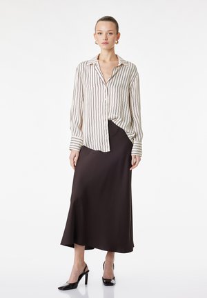 Kvinde iført en hvid og brun lodret striped skjorte med knapper, en mørkebrun lang nederdel og sorte høje hæle, stående mod en hvid baggrund.