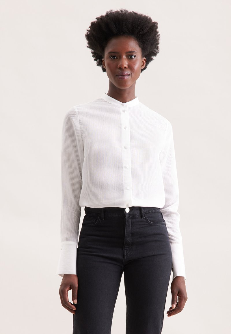 Camicia bianca a maniche lunghe con colletto rialzato e chiusura a bottoni, caratterizzata da sottili righe verticali. Abbinata a jeans neri a vita alta.