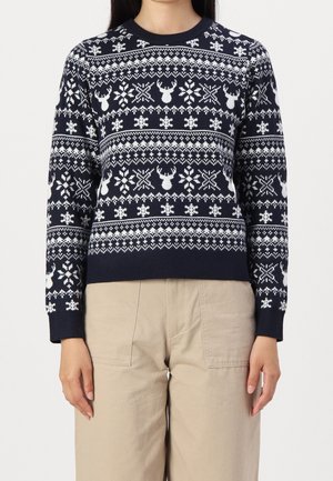 Navy blue sweater with white reindeer and snowflake patterns worn with beige pants.