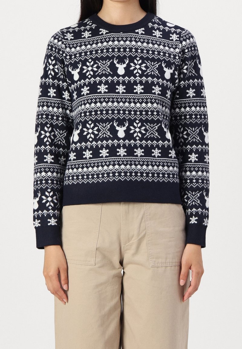 Pull bleu marine avec motifs blancs de rennes et flocons de neige porté avec un pantalon beige.