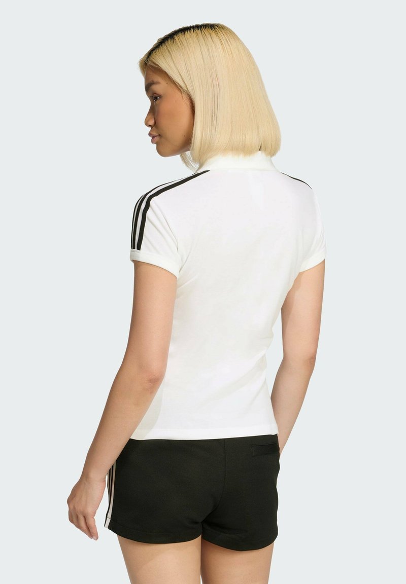 Camisa polo blanca con rayas negras en las mangas, de manga corta y diseño ajustado, combinada con pantalones cortos negros. Tela suave, aspecto casual y deportivo.