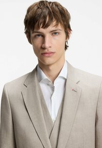 Jeune homme aux cheveux châtain clair portant une veste de costume beige, un gilet assorti et une chemise blanche sur un fond blanc uni.