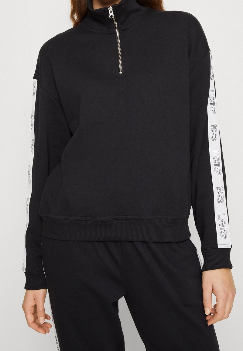 Svart zip-krage sweatshirt i mjukt material, med vit märkes-tape längs ärmarna samt ribbat fåll och ärmslut.