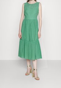Vestido de algodón verde con patrones perforados abiertos, diseño sin mangas y dobladillo en capas. Combinado con sandalias de tacón alto doradas y con tiras.