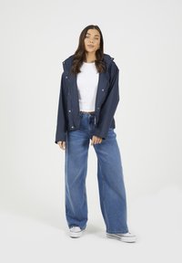 Granatowa wodoodporna kurtka z kapturem, z zapięciami na zatrzaski i elastycznymi mankietami, w połączeniu z jasnoniebieskimi spodniami typu wide-leg i białymi trampkami. Gładkie tło.