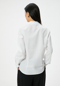 Camisa blanca de manga larga con cuello, textura suave y puños abotonados. La parte trasera presenta un dobladillo curvado y una silueta ajustada.