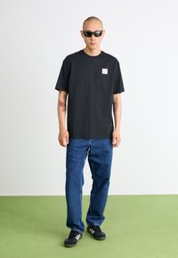 T-shirt en coton noir à manches courtes avec un petit patch graphique, associé à un jean en denim bleu et des baskets noires avec des rayures blanches.
