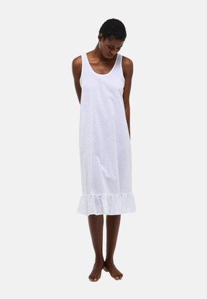 Femme debout pieds nus portant une robe midi blanche sans manches avec de petits pois subtils et un ourlet à volants, regardant vers le bas les mains dans le dos.