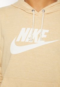 Světle béžová mikina s kapucí a stahovací šňůrkou. Na hrudi se nachází velké bílé logo Nike. Materiál vypadá měkce s jemnou texturou.