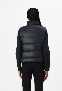 Vesta puffer neagră cu guler ridicat, având secțiuni matlasate și o textură netedă. Asortată cu un top lung cu mâneci întunecat și pantaloni.