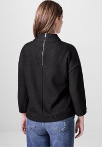 Maglione nero a maglia con colletto alto, caratterizzato da una corta zip sulla schiena, maniche lunghe e dettagli metallici testurizzati.