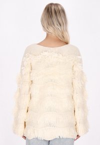 Maglione color crema con un design testurizzato e frangiato. Presenta maniche ampie e una vestibilità ampia, realizzato in tessuto lavorato a maglia.