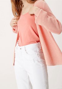 Jasnoróżowy sweter z dzianiny noszony na jasnoróżowej bluzce na ramiączkach, w połączeniu z białymi dżinsami. Sweter ma szerokie rękawy i luźny krój.