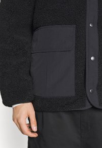 Schwarze Fleecejacke mit einer mattschwarzen Tasche, Druckknöpfen und strukturierten Ärmeln. Das Material ist weich und hat eine zottelige Optik.